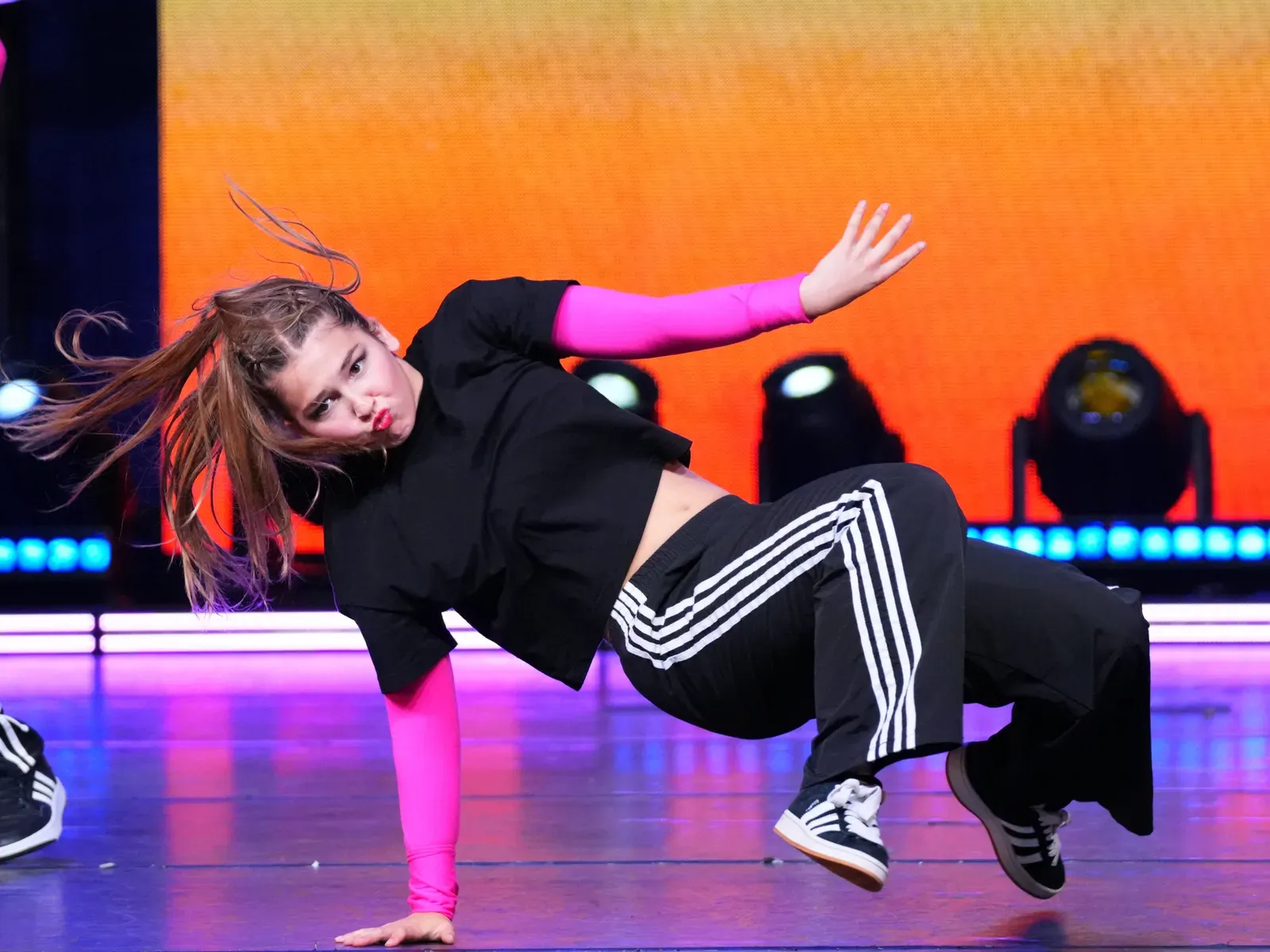A C3 dancer holding a one-handed freeze on stage against an orange backdrop